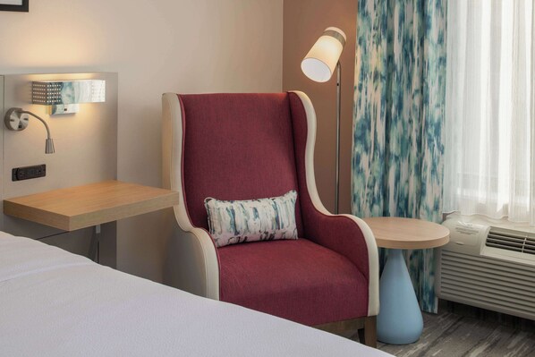 Une chambre d’hôtel avec un fauteuil rouge, une petite table, une table de chevet, une lampe et une fenêtre avec des rideaux.