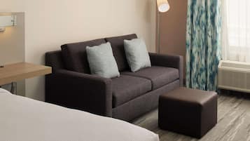Quarto de hotel com sofá marrom escuro, duas almofadas cinzas, um banquinho marrom pequeno, mesa de cabeceira de madeira e uma luminária de chão.