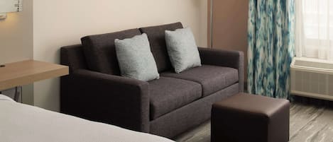 A hotel room with a dark brown sofa, two grey pillows, a small brown ottoman, a wooden side table, and a floor lamp.