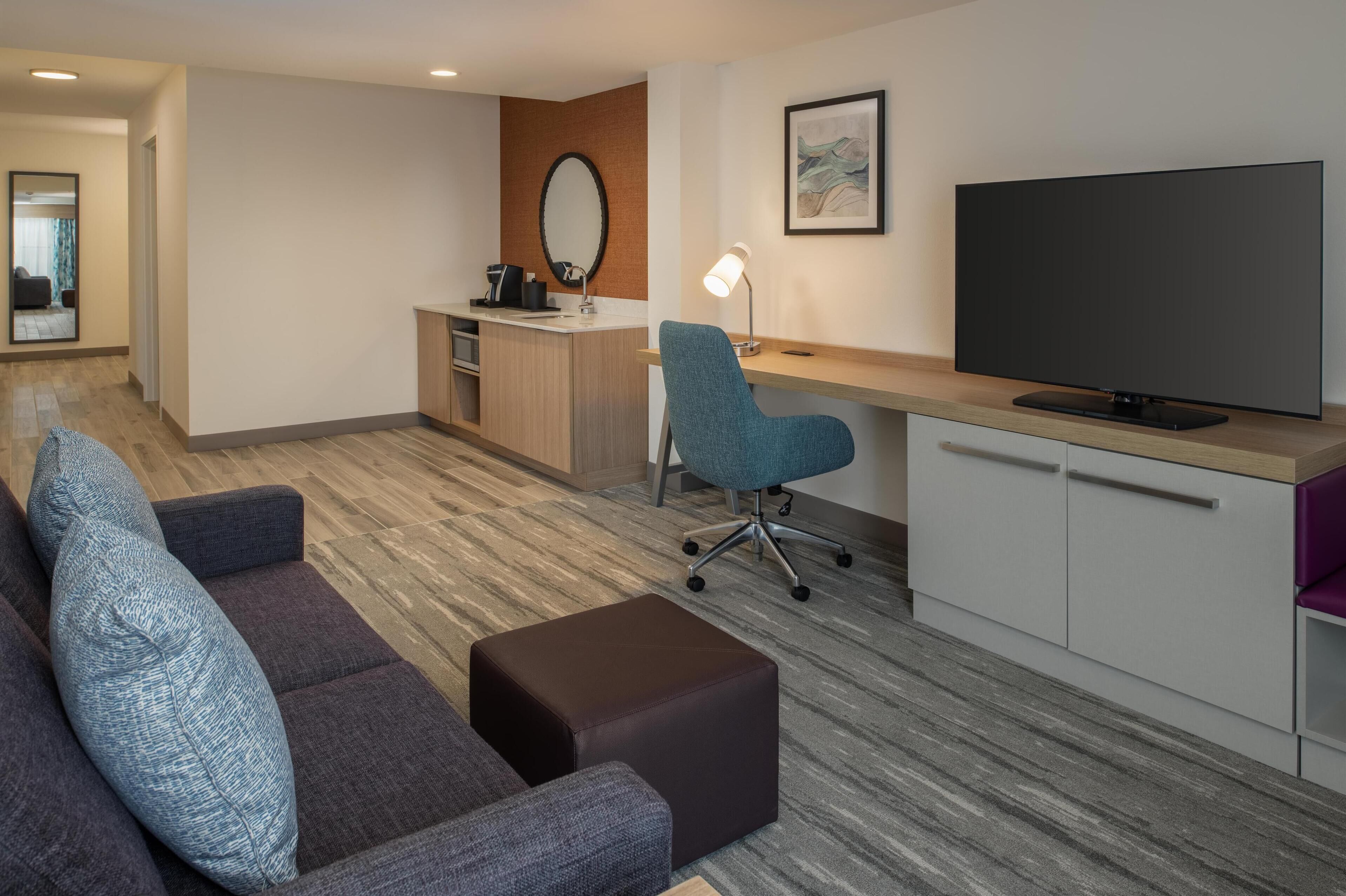 Una habitación de hotel moderna con sofá, reposapiés, escritorio, silla, televisor y una kitchenette pequeña.