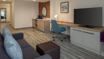 Una habitación de hotel moderna con sofá, reposapiés, escritorio, silla, televisor y una kitchenette pequeña.