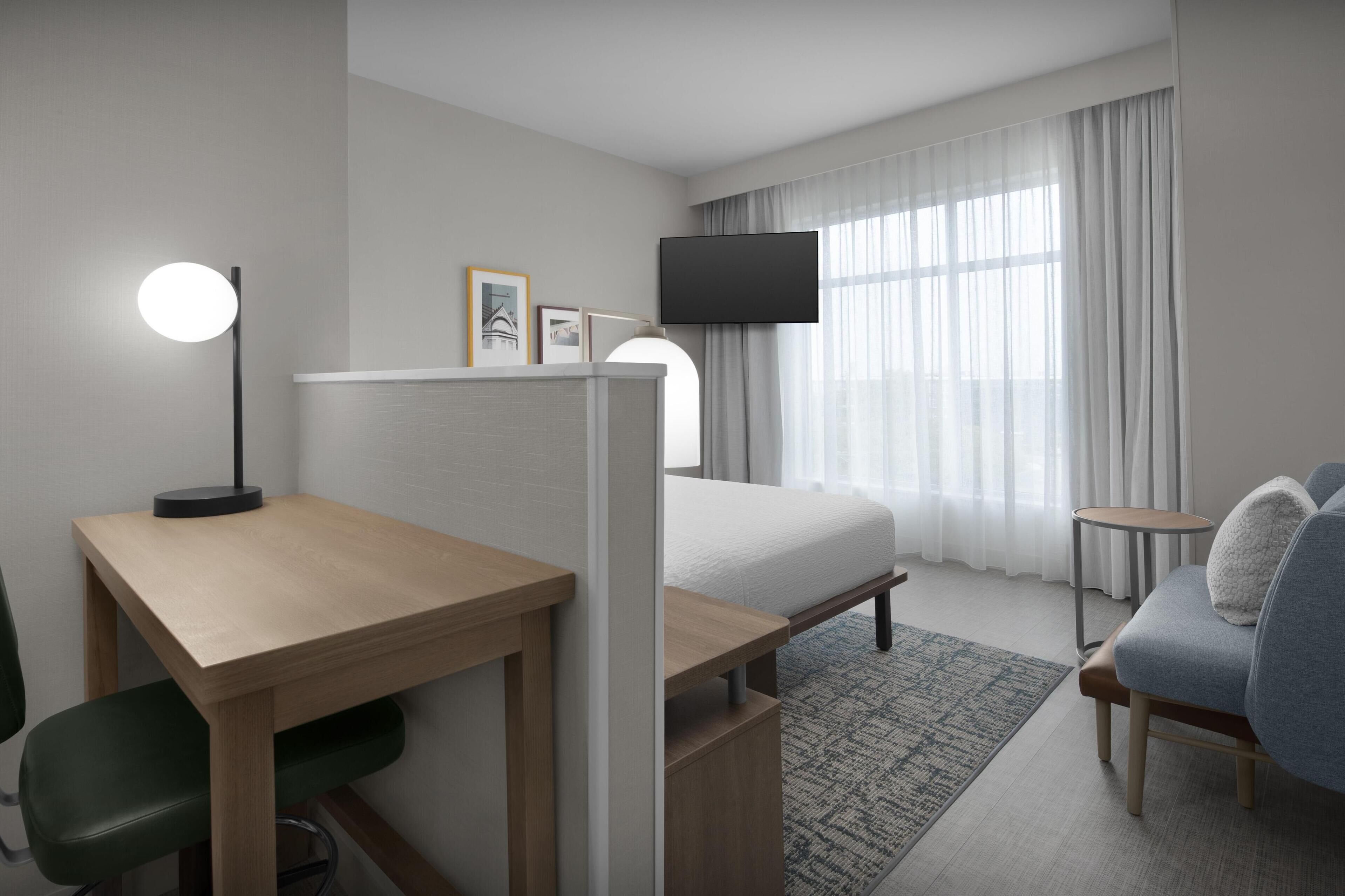 A hotel room with a bed, a desk with a lamp, a TV mounted on the wall, and a sofa.