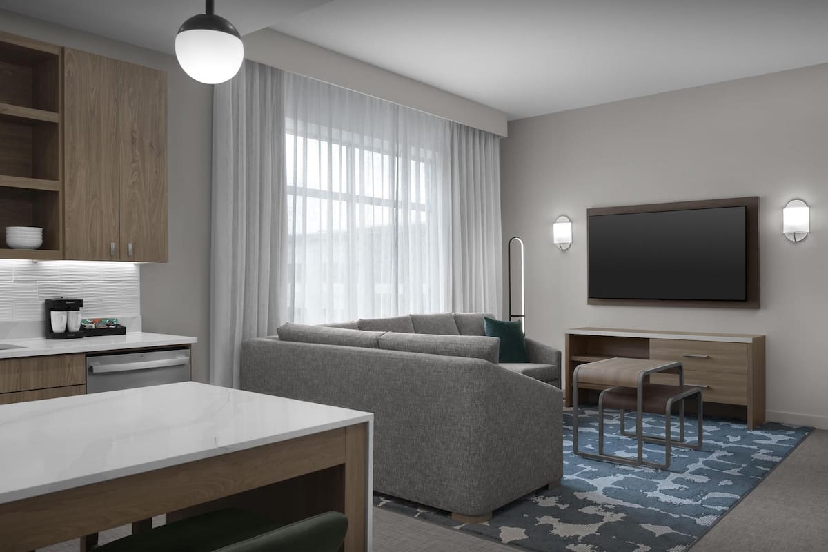 Une chambre d’hôtel moderne équipée d’un canapé, d’une table à manger, d’un téléviseur et d’un coin cuisine avec des placards en bois et une machine à café.