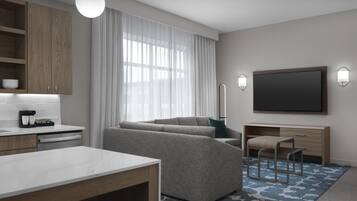 A modern hotel room with a sofa, a dining table, a TV, and a kitchen area with wooden cabinets and a coffee machine.