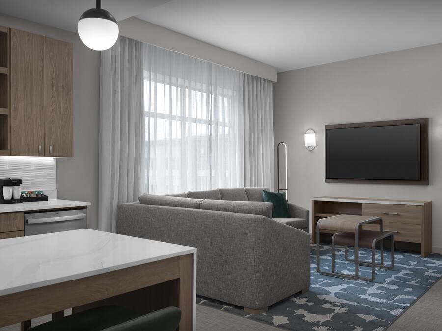 A modern hotel room with a sofa, a dining table, a TV, and a kitchen area with wooden cabinets and a coffee machine.