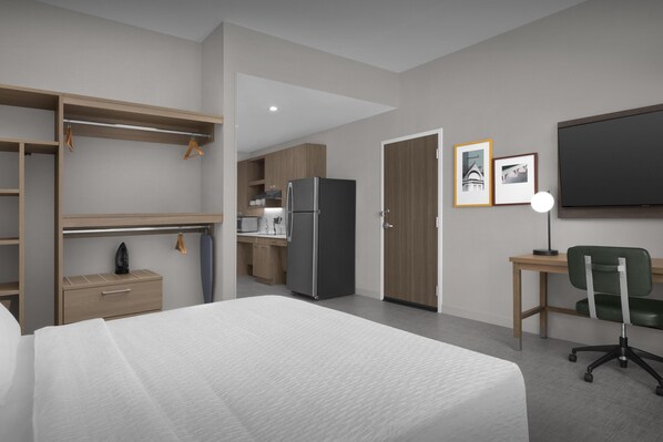 A modern hotel room with a bed, a desk with a chair, a kitchenette, a wardrobe, and a TV.