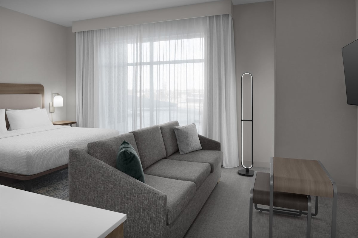 Une chambre d’hôtel moderne avec un lit, un canapé gris, une petite table et une lampe sur pied.