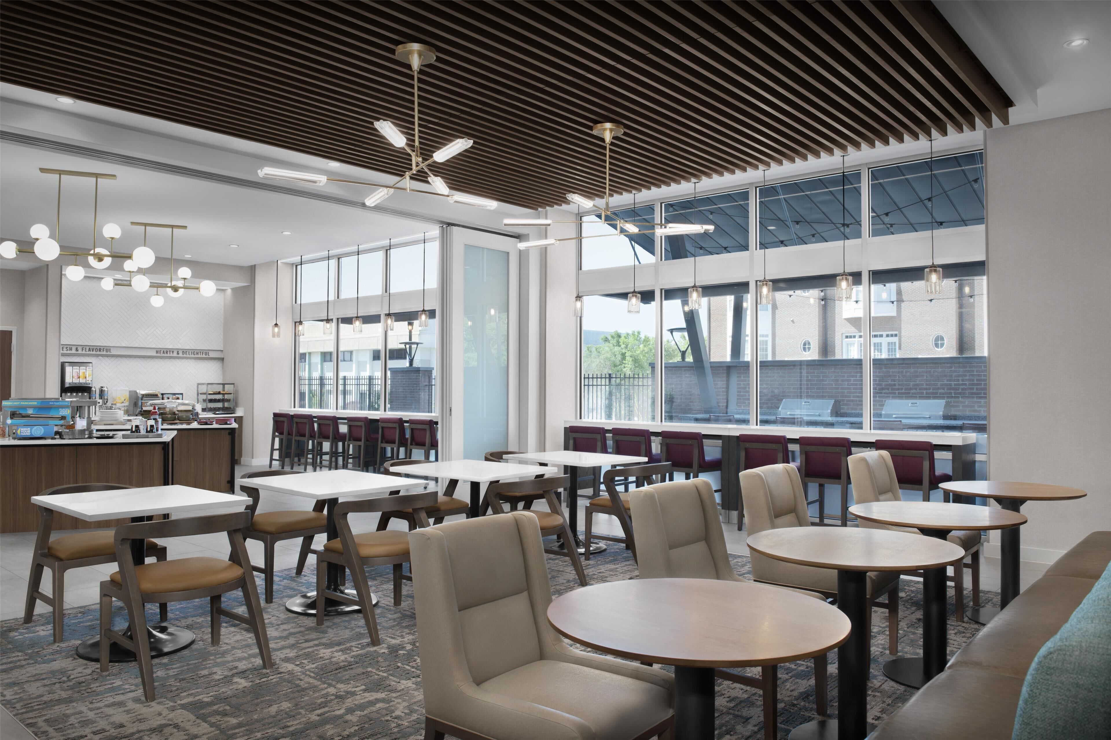Un espace de restauration moderne, avec des tables et des chaises, de grandes fenêtres et une cuisine en arrière-plan.