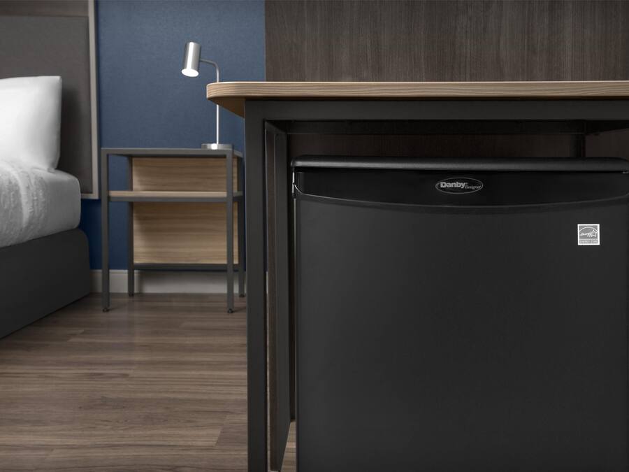 Ein kleiner Kühlschrank in einem Hotelzimmer mit Bett, Nachttisch und Wandlampe.