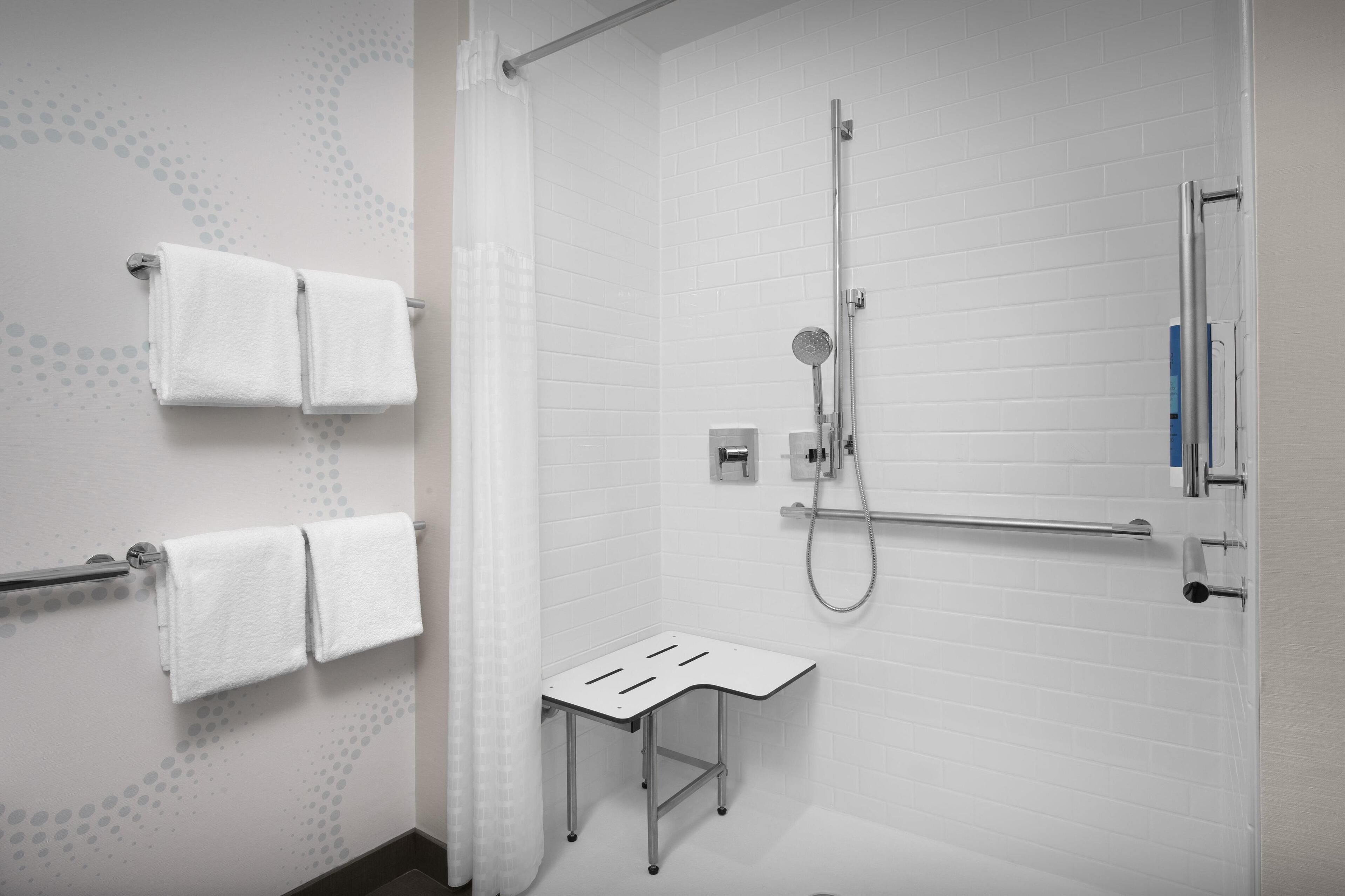 Une salle de bain moderne avec des murs recouverts de carreaux blancs, une douche équipée d’un pommeau de douche à main, un porte-serviettes avec deux serviettes et un petit marchepied.
