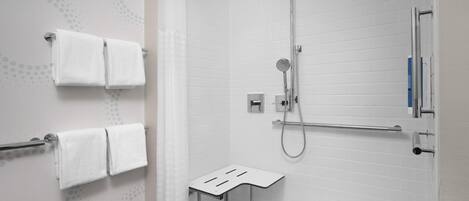 A modern bathroom with white tiled walls, a shower with a handheld showerhead, a towel rack with two towels, and a small step stool.