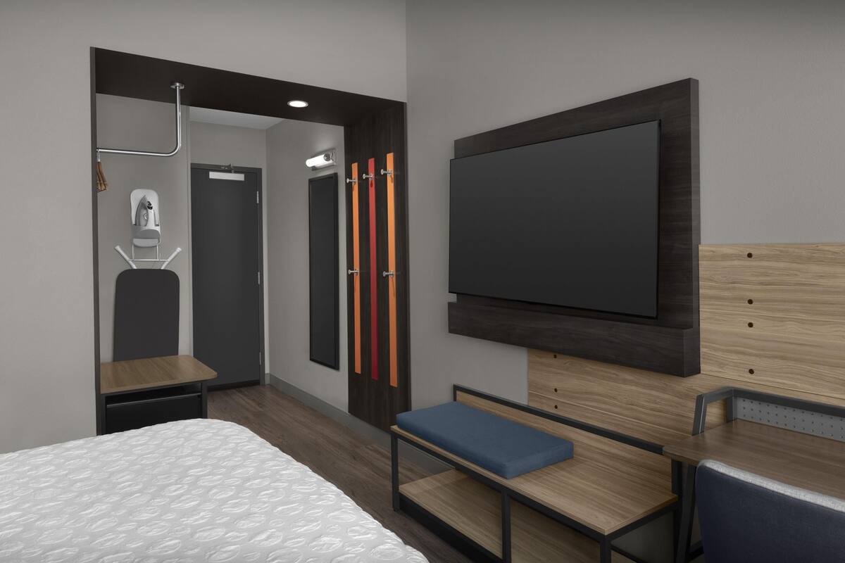 Una habitación de hotel moderna con una cama, un escritorio, un banco, un televisor montado en la pared y un armario con espejo.