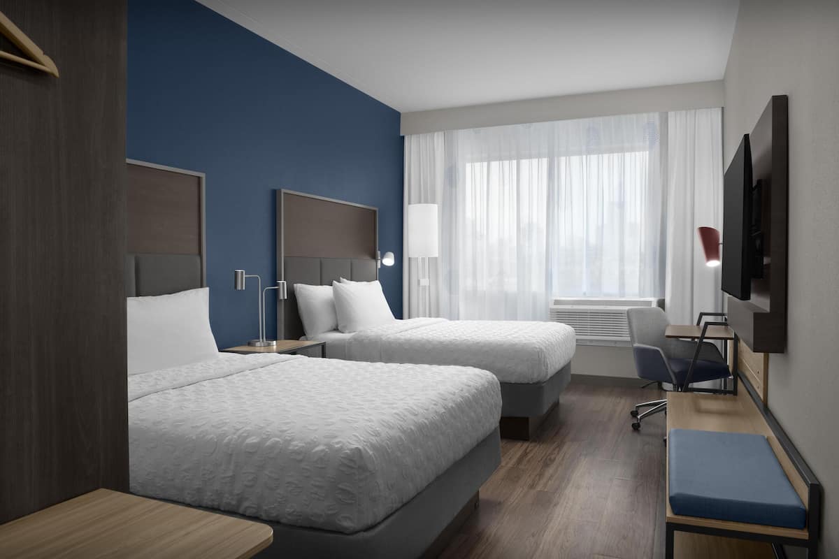 Una habitación de hotel con dos camas, un escritorio, una silla, un televisor y una ventana con cortinas.