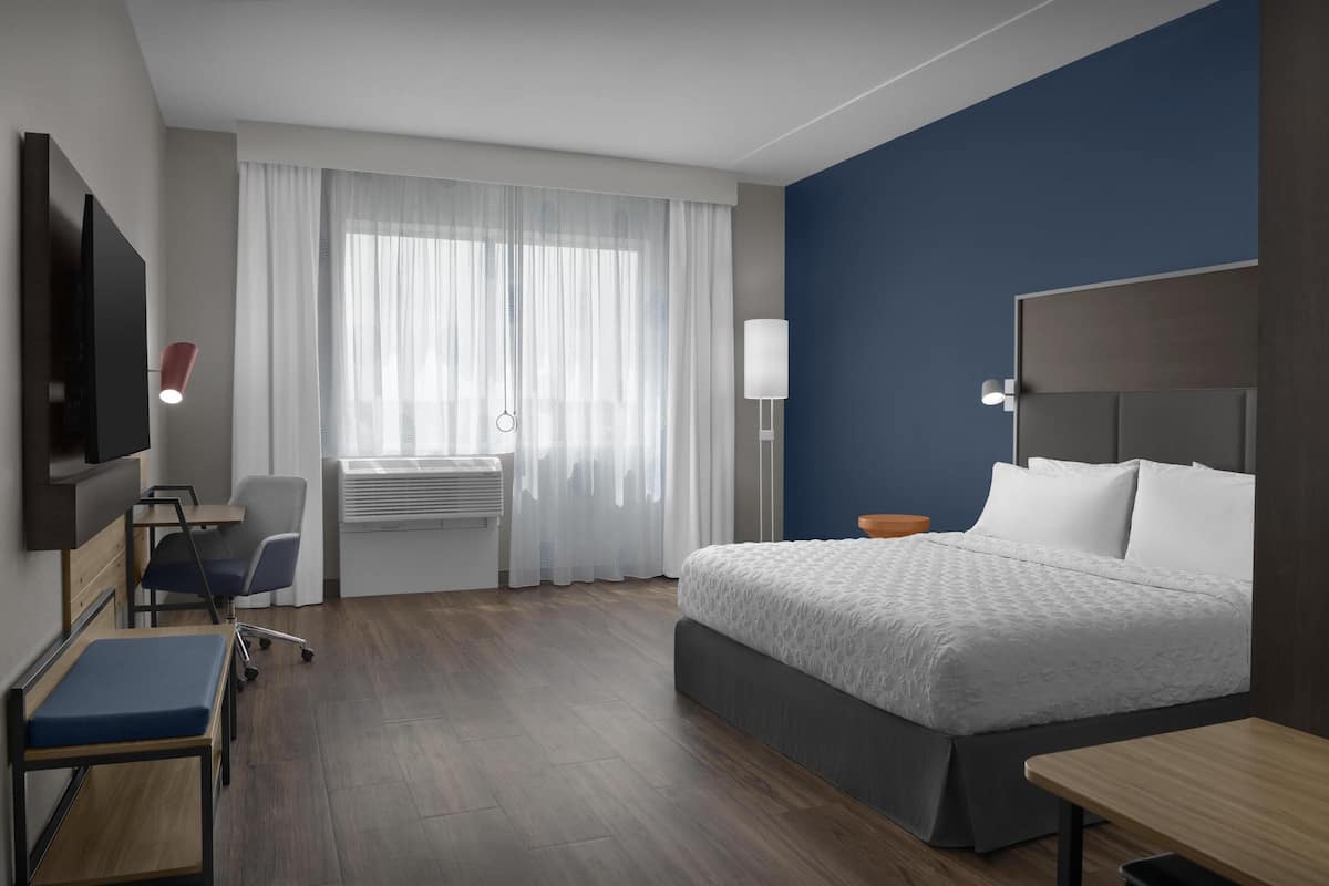 Habitación de hotel con cama, escritorio, silla, televisor y ventana con cortinas.