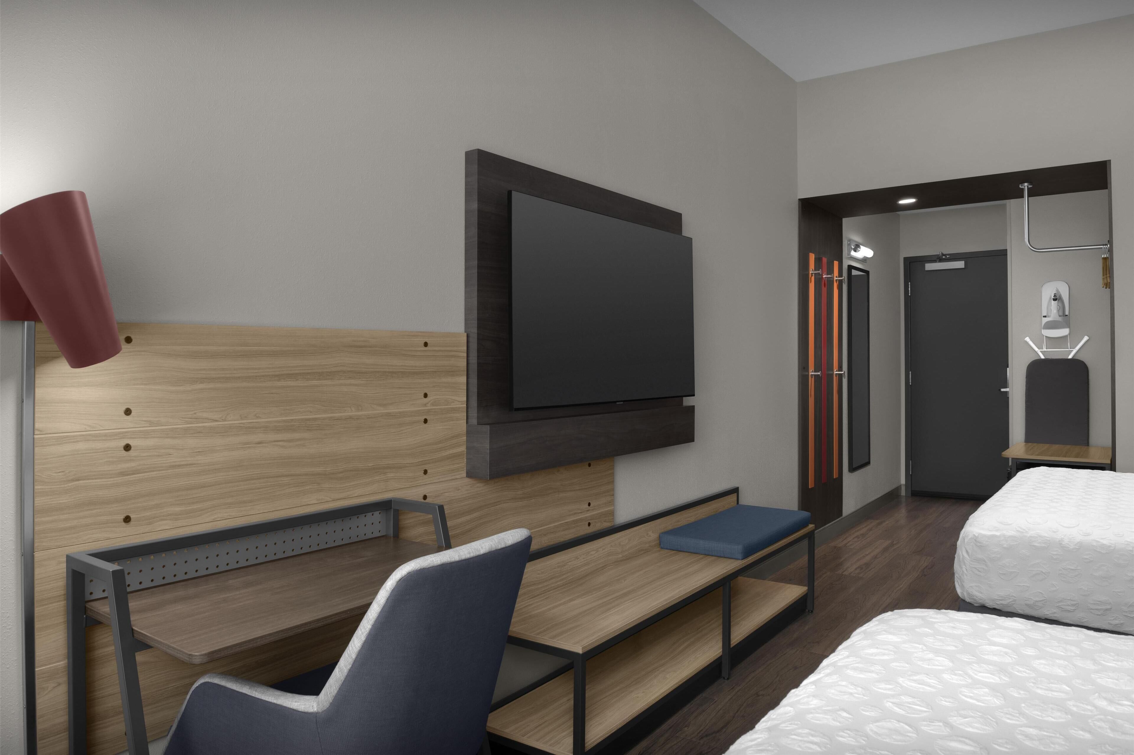 A modern hotel room with a wooden headboard, a flat-screen TV mounted on the wall, a desk with a chair, a bed with white bedding, and a small seating area.