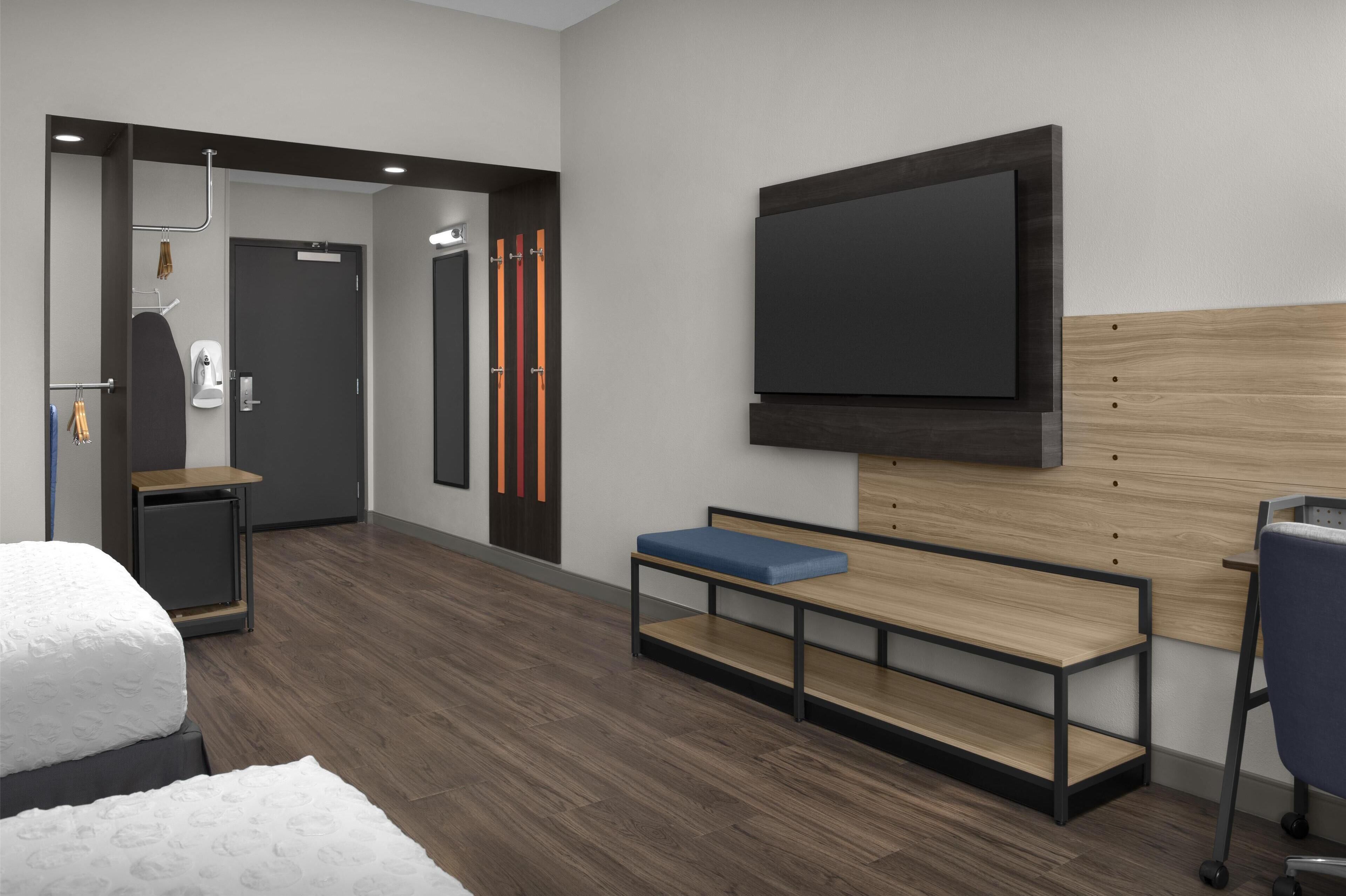 Een moderne hotelkamer met een bed, een bureau, een bankje, een aan de muur bevestigde televisie en een deur met een handdoekhouder.
