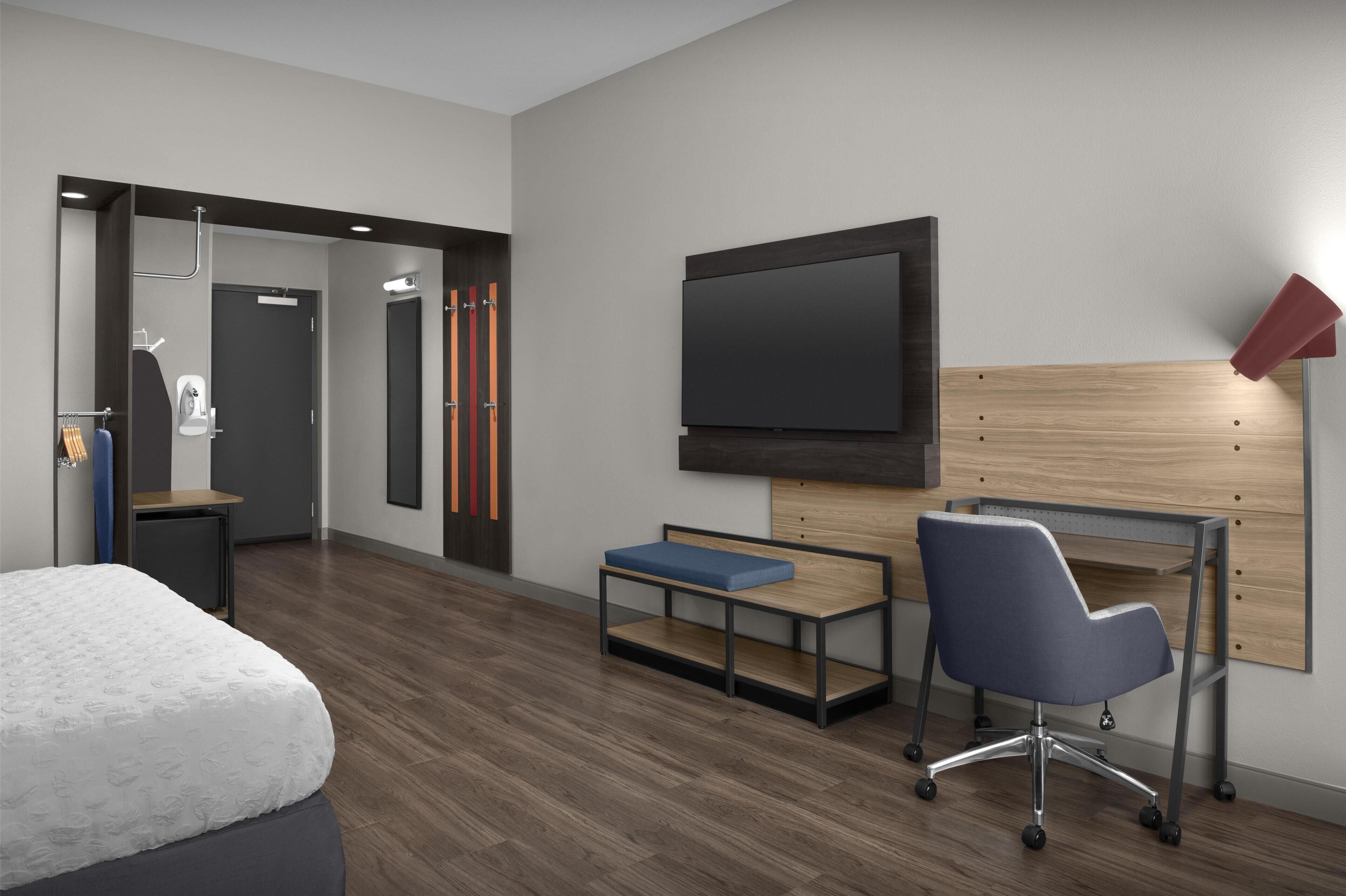 A modern hotel room with a bed, a desk with a chair, a TV mounted on the wall, and a shower area.