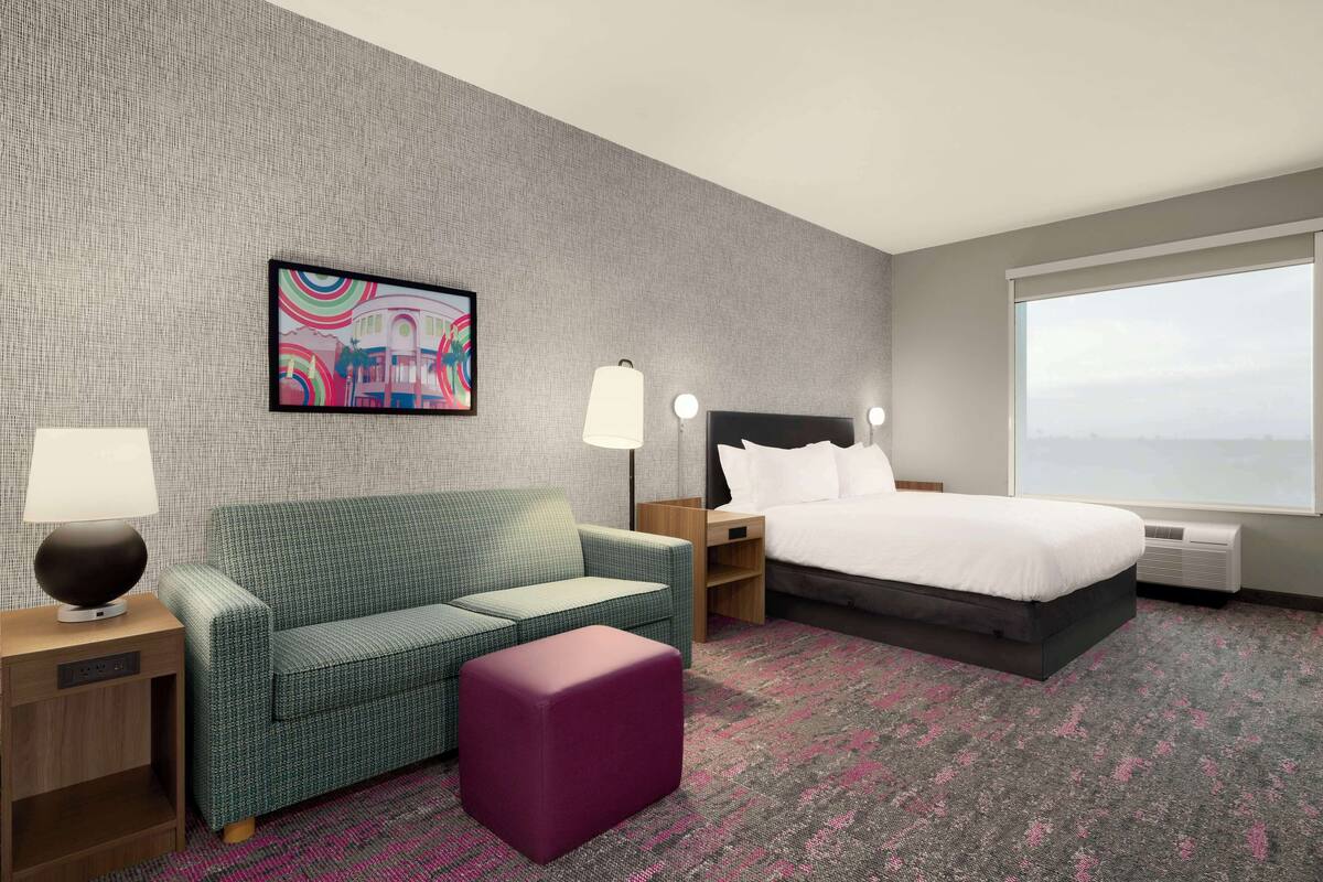 Ein Hotelzimmer mit einem grünen Sofa, einem violetten Hocker, einem Bett mit weißer Bettwäsche, einem Nachttisch mit Lampe, einem an der Wand montierten Fernseher und einem großen Fenster mit Blick.