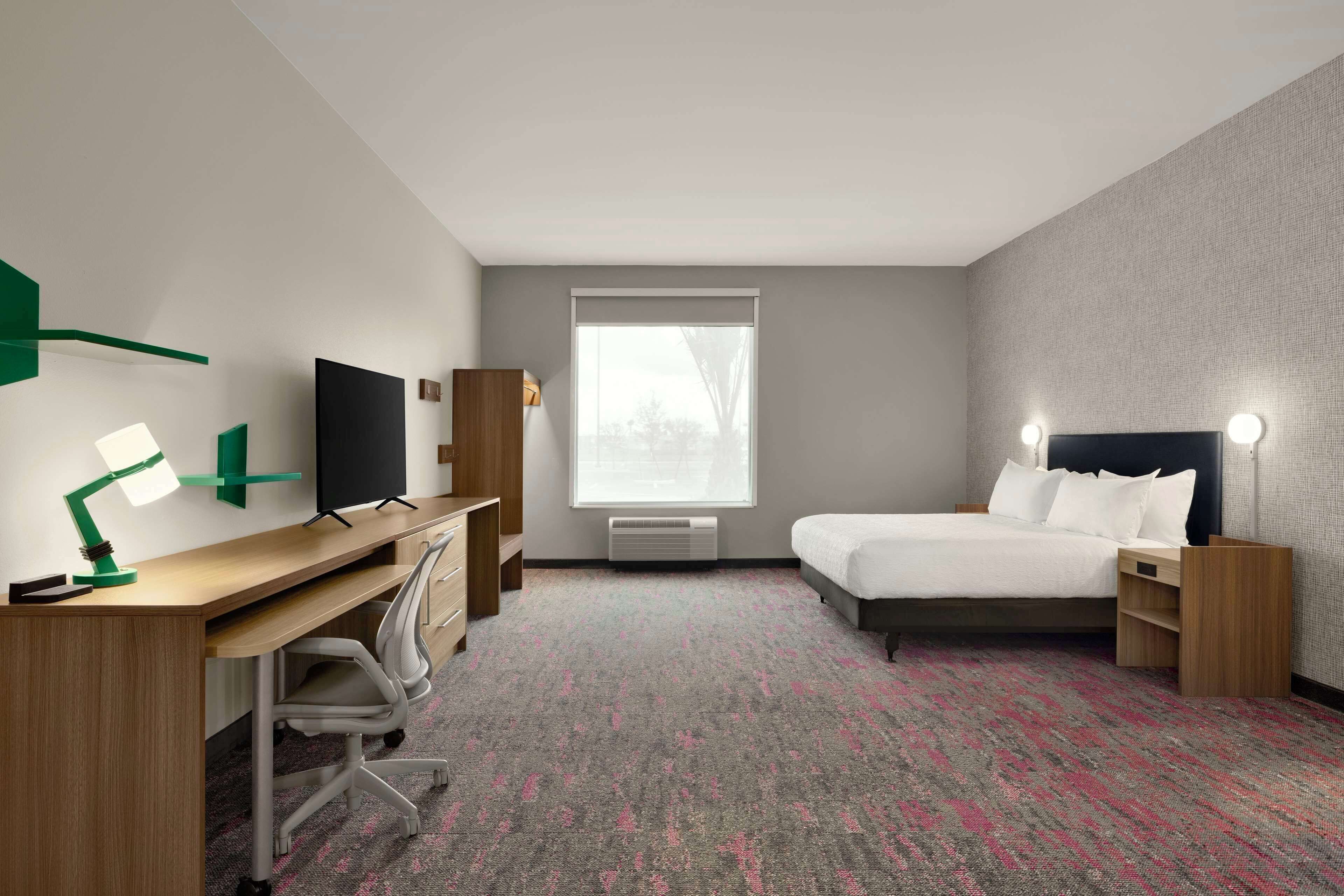 A hotel room with a desk, a bed, a TV, a window, and a carpet.
