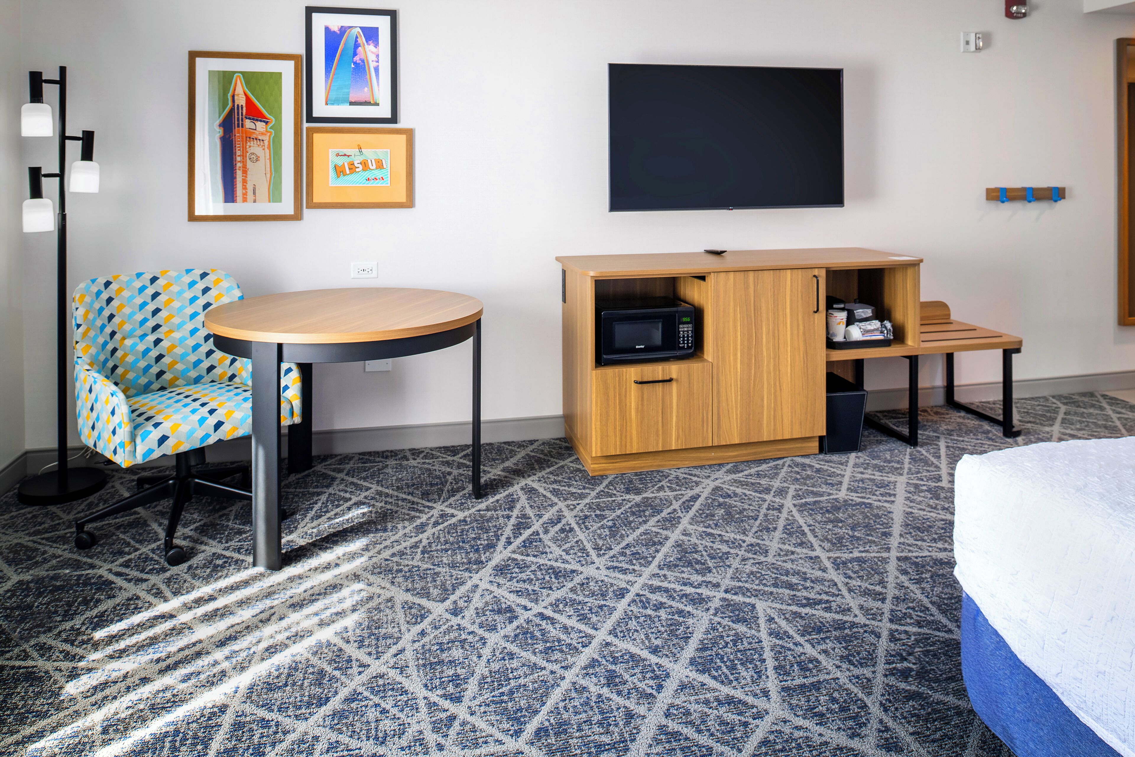 A hotel room with a patterned carpet, a round table, a blue and yellow patterned chair, a wooden cabinet with a microwave, and a bed with a white mattress.