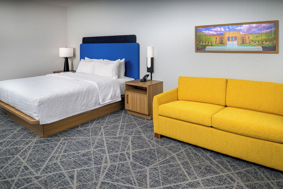 Habitación de hotel con cama, mesita de noche, lámpara, teléfono, sofá amarillo y un cuadro en la pared.