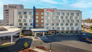 Un hôtel moderne, reconnaissable à son grand panneau Hilton, à son panneau Hampton Inn by Hilton, et doté d’un parking avec une zone abritée et des parterres aménagés.