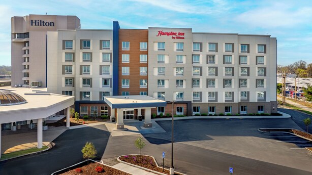 A modern hotel with a prominent Hilton sign, a Hampton Inn by Hilton sign, and a parking lot with a covered area and landscaped beds. - Hampton Inn By Hilton St Louis Airport (St. Louis)