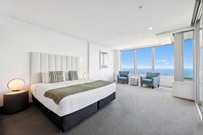 A modern hotel room with a large bed, a seating area with two chairs, a small table, and a large window offering a view of the ocean. - Q1 Resort & Spa - Official (Surfers Paradise)