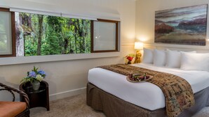 Habitación de hotel con una cama grande, una ventana con vistas a un bosque, un cuadro en la pared y una mesita con flores.