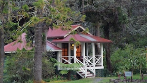 A small, rustic cabin with a red roof, surrounded by lush greenery and trees. - Private Cottage at Boutique Hotel in Volcano Village (Volcano)
