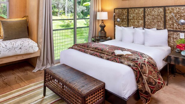A hotel room with a large bed, a bench, a nightstand, and a view of a garden through a glass door. - Eclectic Bungalow close to Volcanoes National Park (Volcano)