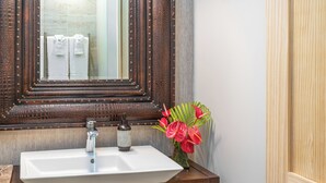 A bathroom with a white sink, a mirror with a wooden frame, and a vase with red flowers. - Eclectic Bungalow close to Volcanoes National Park (Volcano)