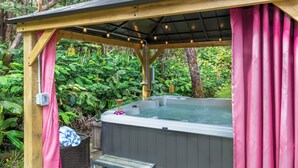 A hot tub under a wooden pergola with pink curtains, surrounded by lush greenery. - Eclectic Bungalow close to Volcanoes National Park (Volcano)