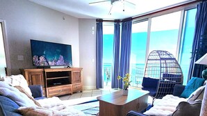 A modern hotel room with a large bed, a sofa, a TV, a ceiling fan, and a balcony with a view of the ocean. - AQUA 1509 — Luxury Oceanfront Condo• 3 BD• Steps to Pier Park (Panama City Beach)
