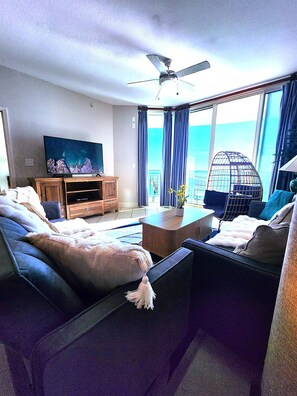 A modern hotel room with a large bed, a sofa, a TV, a ceiling fan, and a balcony with a view of the ocean. - AQUA 1509 — Luxury Oceanfront Condo• 3 BD• Steps to Pier Park (Panama City Beach)