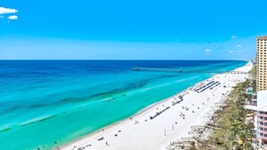 A beach with clear turquoise waters, a pier extending into the sea, and a row of beachside buildings. - AQUA 1509 — Luxury Oceanfront Condo• 3 BD• Steps to Pier Park (Panama City Beach)