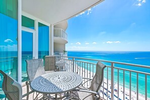 Un balcon avec une table ronde et des chaises, donnant sur une plage aux eaux d’un bleu limpide et sous un ciel radieux.