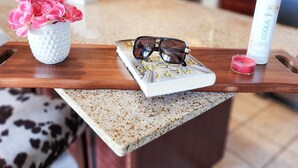 Un comptoir de cuisine orné d’un vase blanc contenant des fleurs roses, d’une paire de lunettes de soleil, d’un livre, d’une petite bougie et d’une bouteille de produit de bain minéral de la marque COOLA.