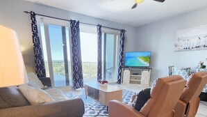 A spacious living room with a sofa, armchairs, a coffee table, and a TV. There is a ceiling fan and a balcony with curtains. - On the most private end of the building, accessible and gorgeous! (Panama City Beach)