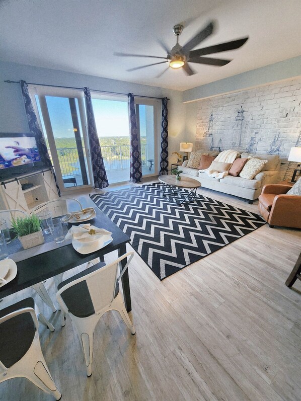 A modern living room with a large window, a ceiling fan, a dining area with a table and chairs, and a sofa with decorative pillows. - Immaculate! Large and inviting! Dive into the best vacation NOW! (Panama City Beach)