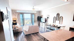 A modern kitchen with a large island, a dining area with a table and chairs, a sofa, and a view of the outdoors through large windows.