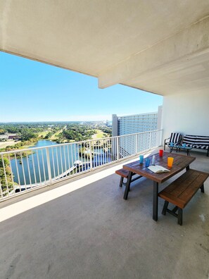 A balcony with a view of a lake and buildings, a wooden table with drinks, and a striped bench. - Exceptional unit! Extra large offering the best set up and views! (Panama City Beach)