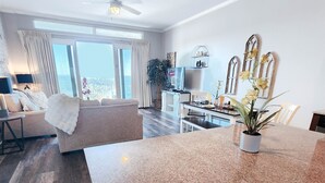 A modern living room with a large window, a ceiling fan, a dining area with a granite countertop, and a kitchen with wooden cabinets.
