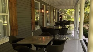 An outdoor covered patio with multiple tables and chairs, wooden columns, and a view of the surrounding greenery.