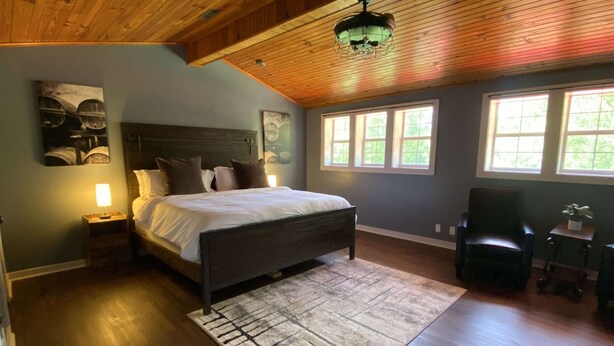 A bedroom with a wooden ceiling, a bed with a headboard, two bedside tables with lamps, a rug, and a chair.