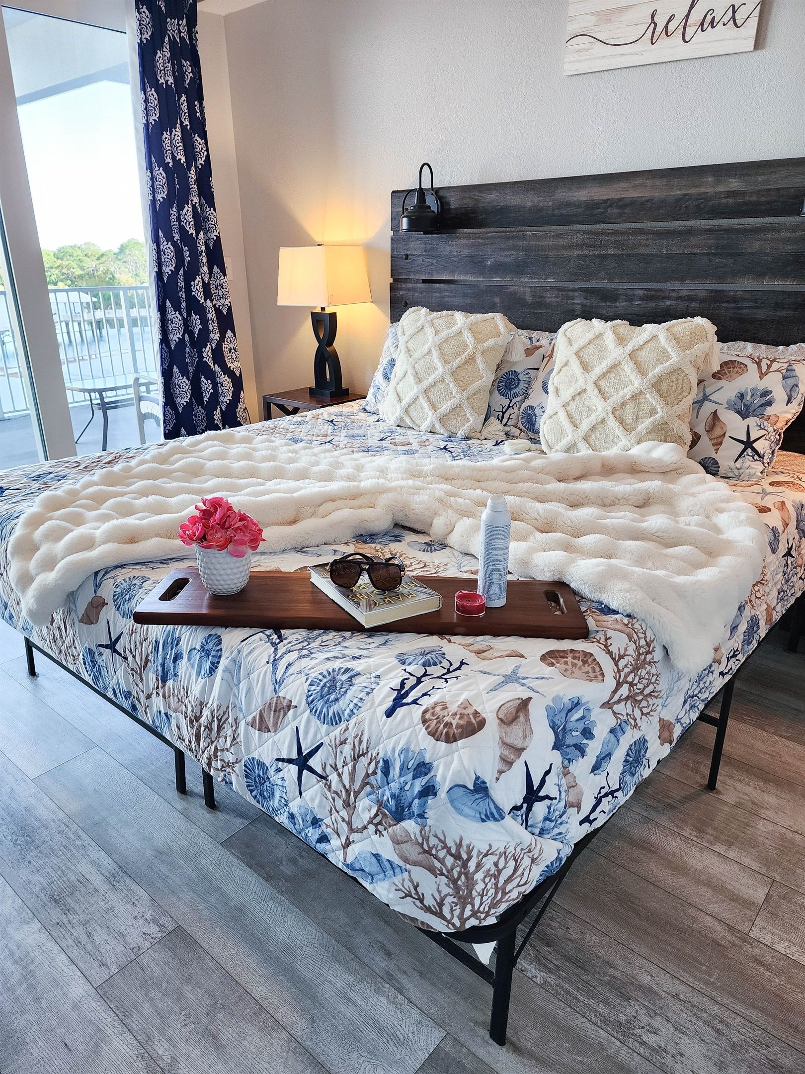 A neatly made bed with a floral-patterned duvet, a decorative pillow, and a bedside table with a lamp, book, and a small vase.
