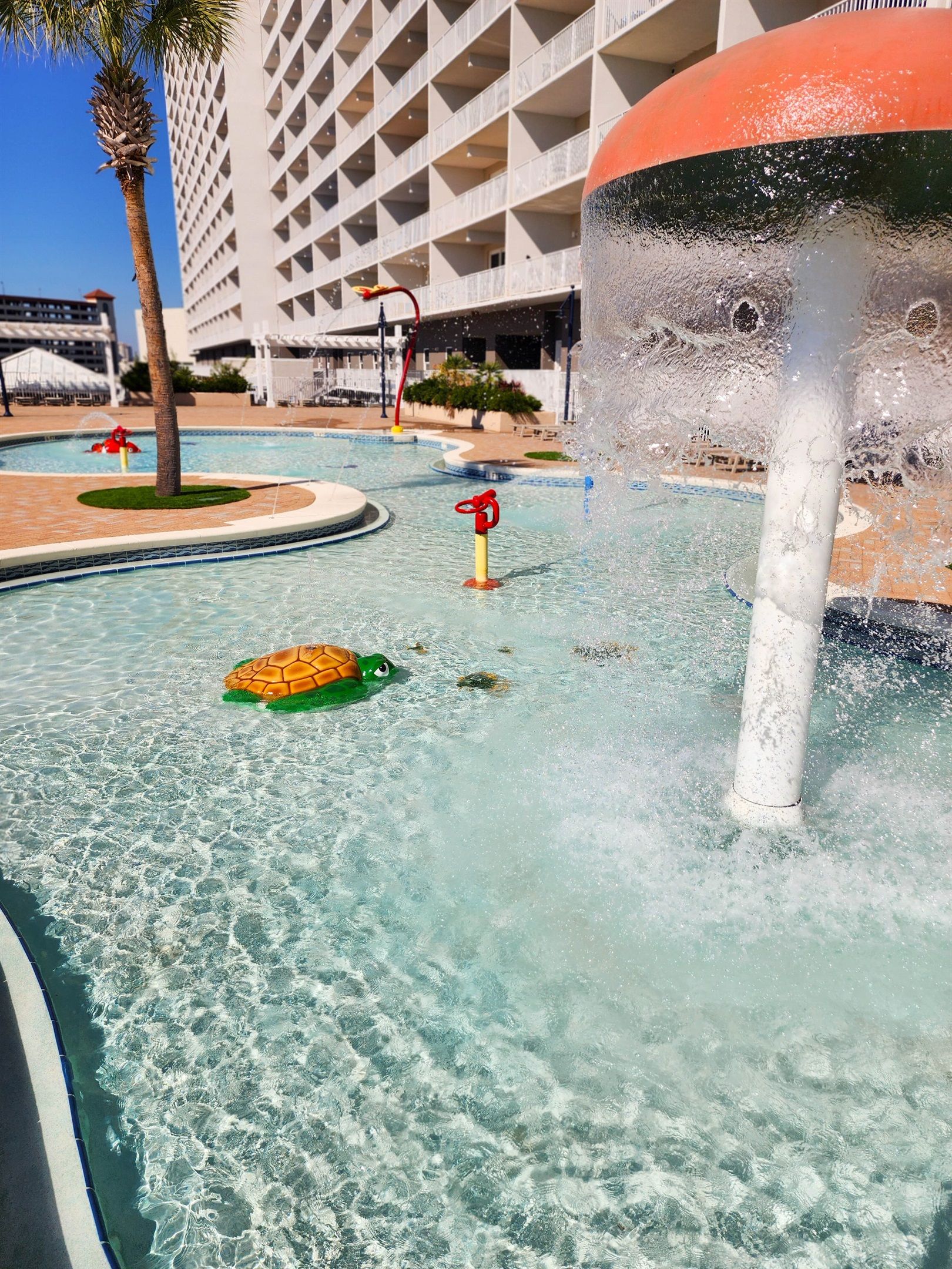 En swimmingpool med et springvand, palmer og en bygning i baggrunden.