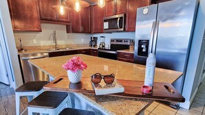 A modern kitchen with wooden cabinets, a granite countertop, a stainless steel refrigerator, and a built-in oven. There are bar stools and a small island with a vase of flowers.