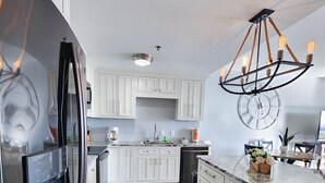A modern kitchen with white cabinetry, a stainless steel refrigerator, a marble countertop, and a unique chandelier. - Exceptional unit 633!Accessible and  Inviting! Perfect Beach set up! (Panama City Beach)
