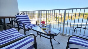 A balcony with striped lounge chairs, a small table with a book and bottles, and a view of a body of water and distant landscape. - Gorgeous Unit, Fantastic Location, Best amenities with a private boardwalk (Panama City Beach)